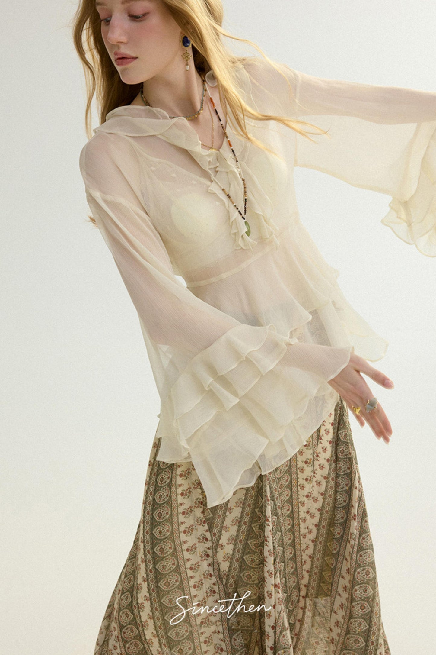 Woman wearing a sheer beige blouse with long sleeves and a patterned skirt on a white background.