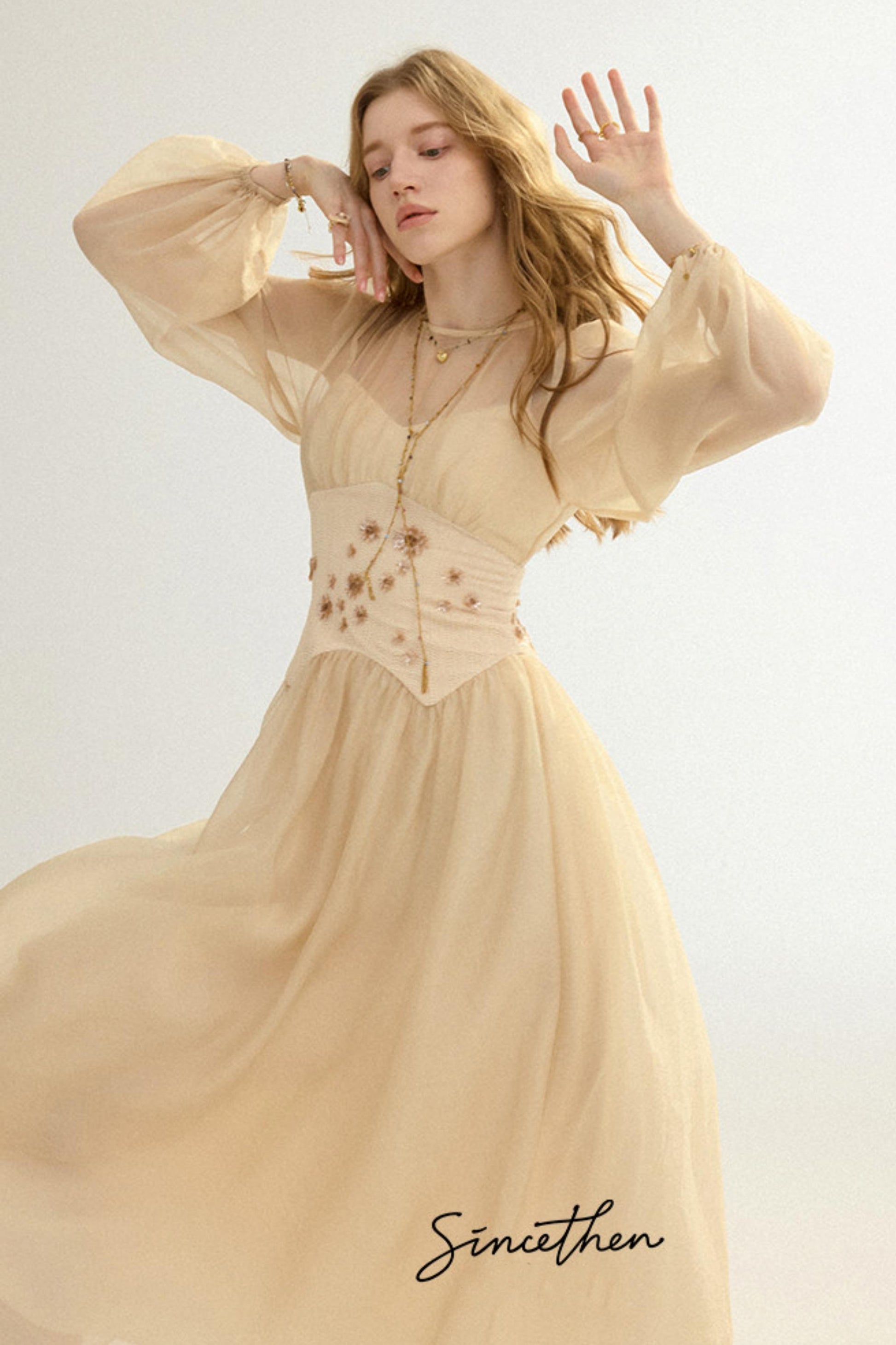 Woman wearing a beige dress with floral details on a light background, with 'Sineithen' branding.