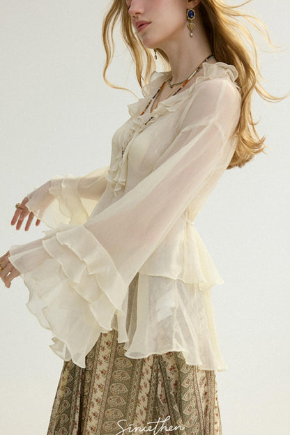 Woman wearing a sheer beige blouse with long sleeves and a patterned skirt on a white background.