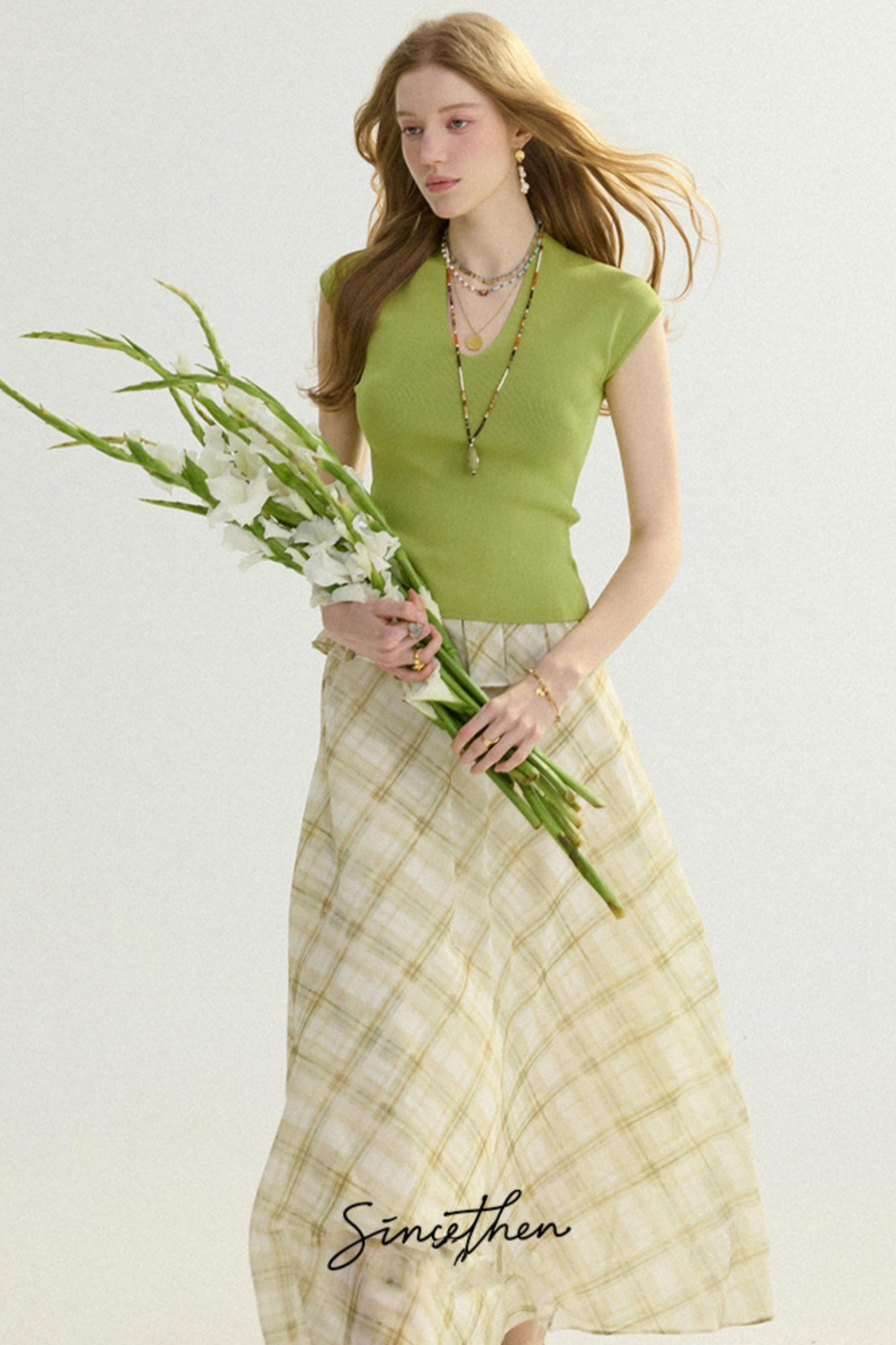 Woman wearing a green top and beige plaid skirt holding flowers against a white background