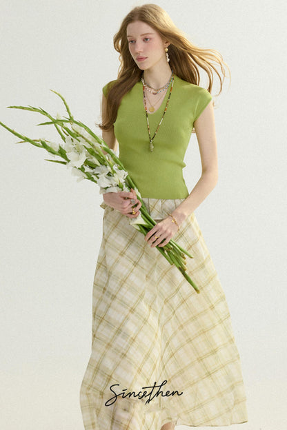Woman wearing a green top and beige plaid skirt holding flowers against a white background
