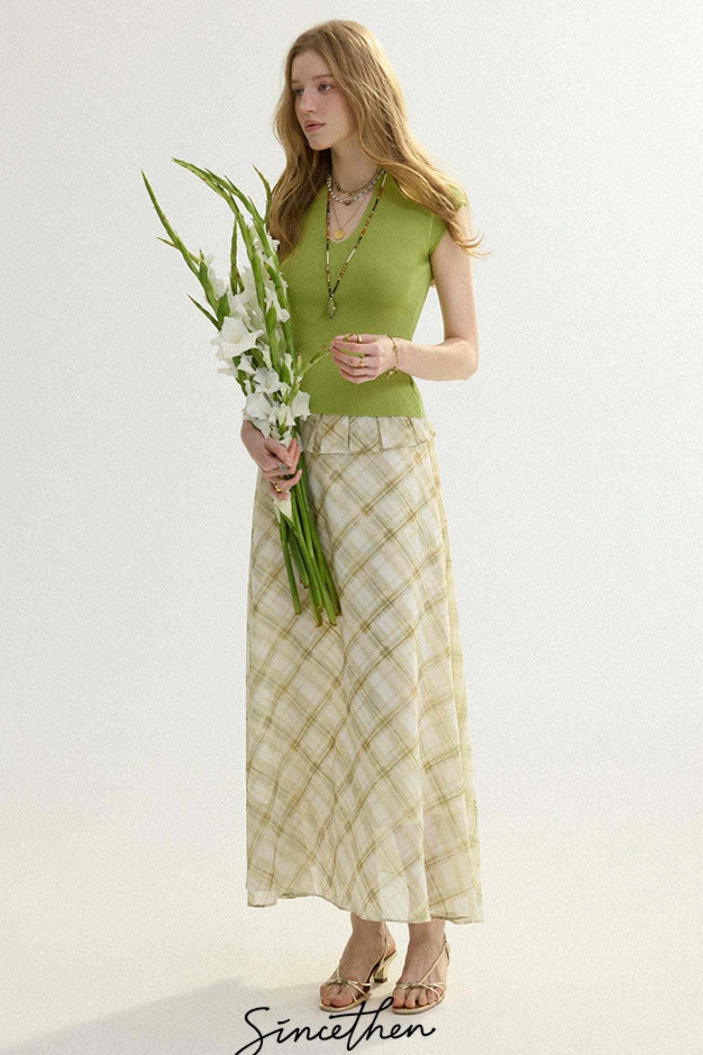 Woman in a green top and patterned skirt holding flowers against a white background