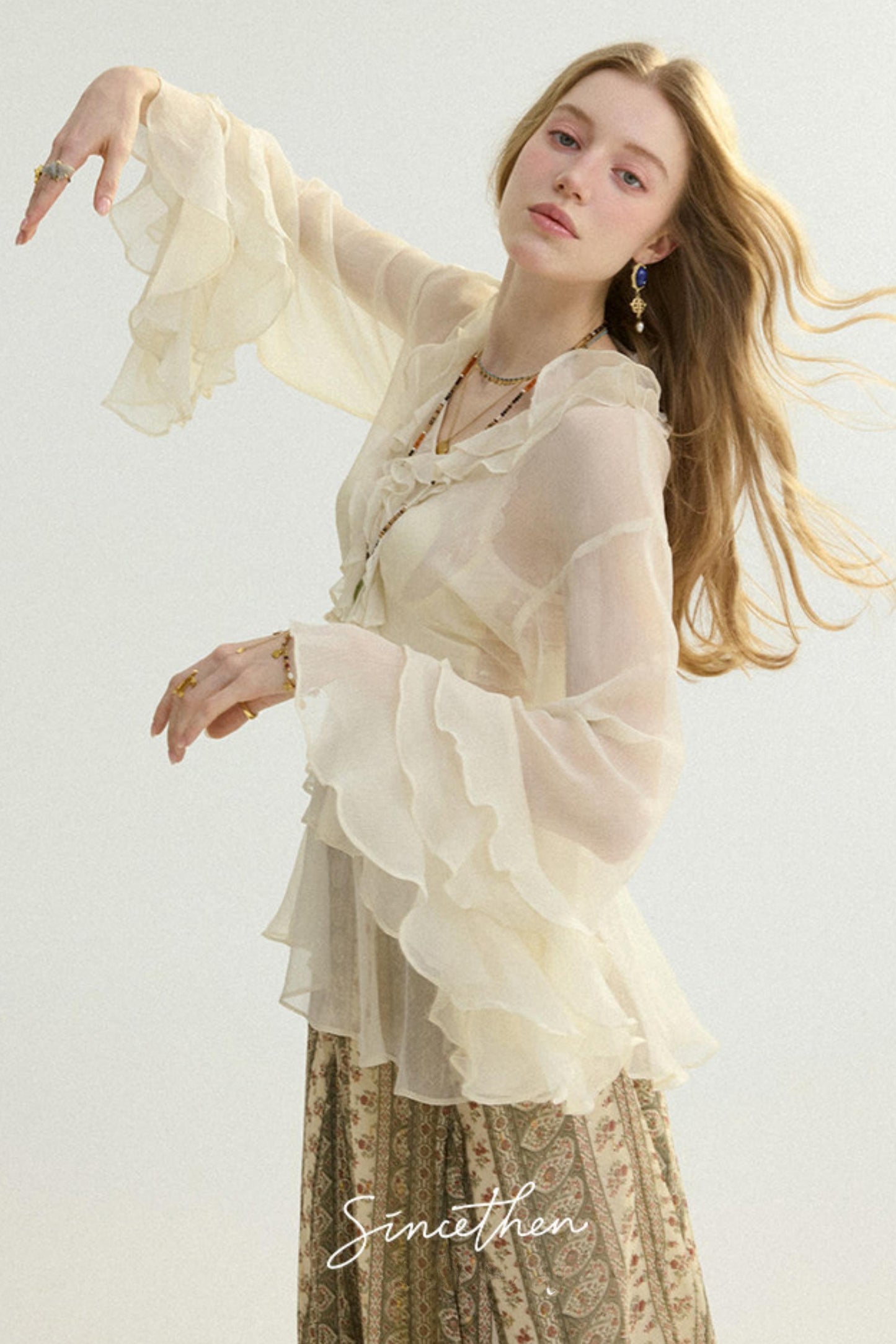 Woman wearing a sheer beige blouse with ruffled sleeves and a patterned skirt on a light background.
