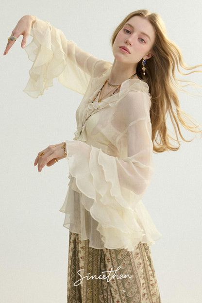 Woman wearing a sheer beige blouse with ruffled sleeves and a patterned skirt on a light background.