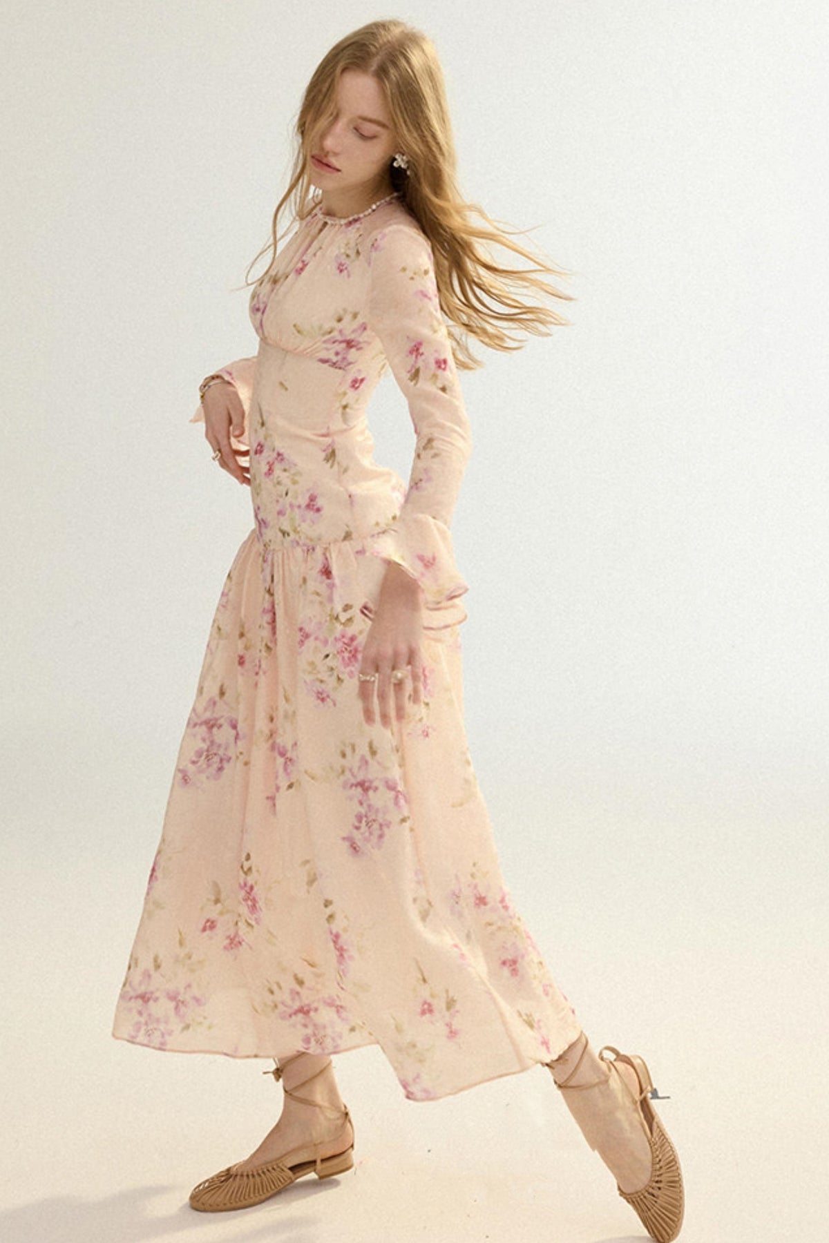 A woman modeling a long-sleeved, midi-length floral dress in a pale pink color with pink and green floral patterns. She is wearing beige lace-up flat shoes against a plain, light off-white studio background.