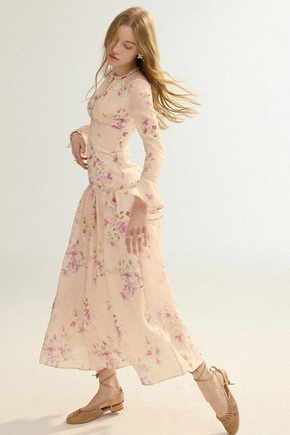 A woman modeling a long-sleeved, midi-length floral dress in a pale pink color with pink and green floral patterns. She is wearing beige lace-up flat shoes against a plain, light off-white studio background.