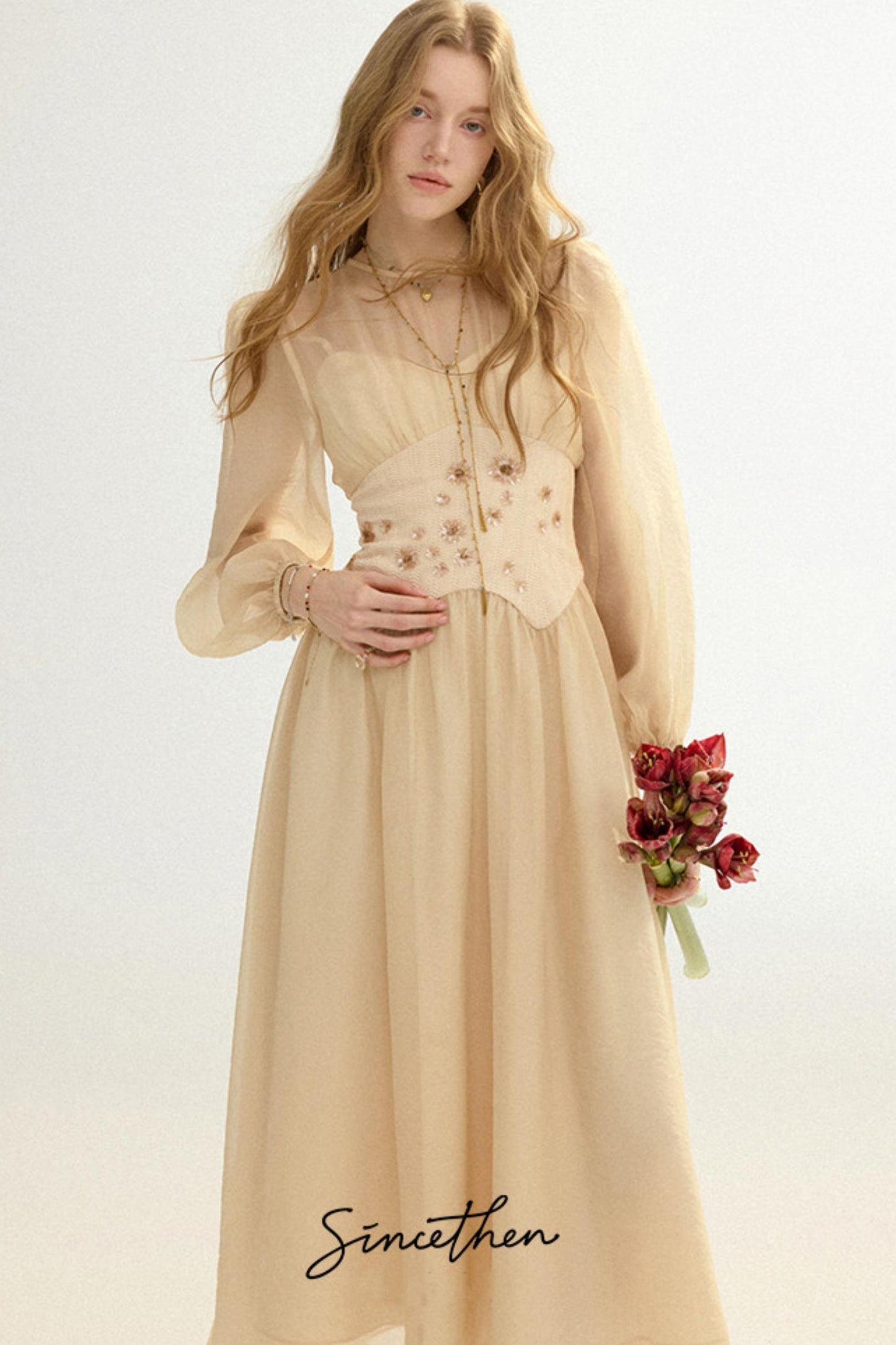 Woman wearing a beige dress with floral details, holding flowers against a white background.