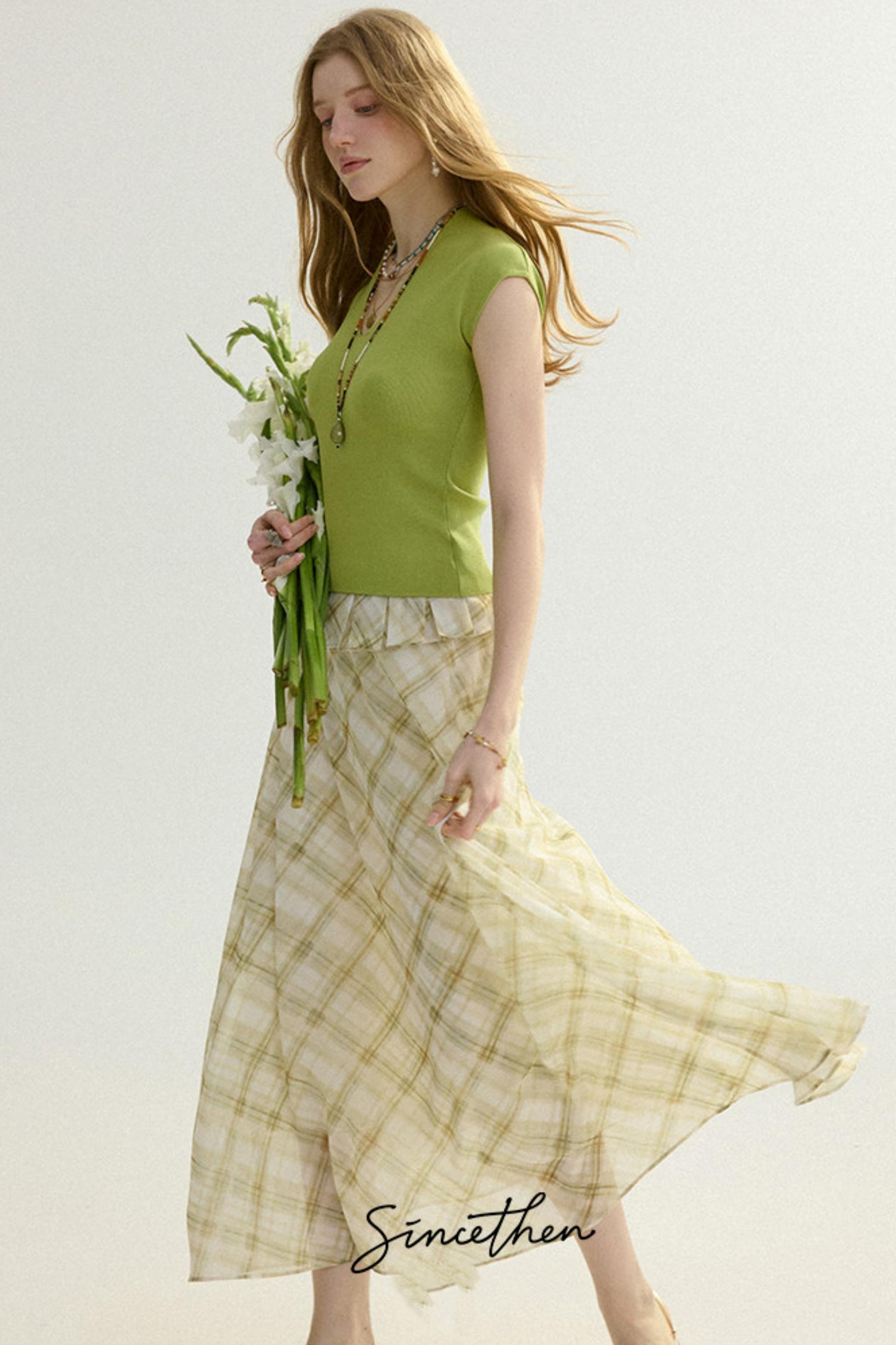 Woman wearing a green sleeveless top and plaid skirt holding flowers against a light background