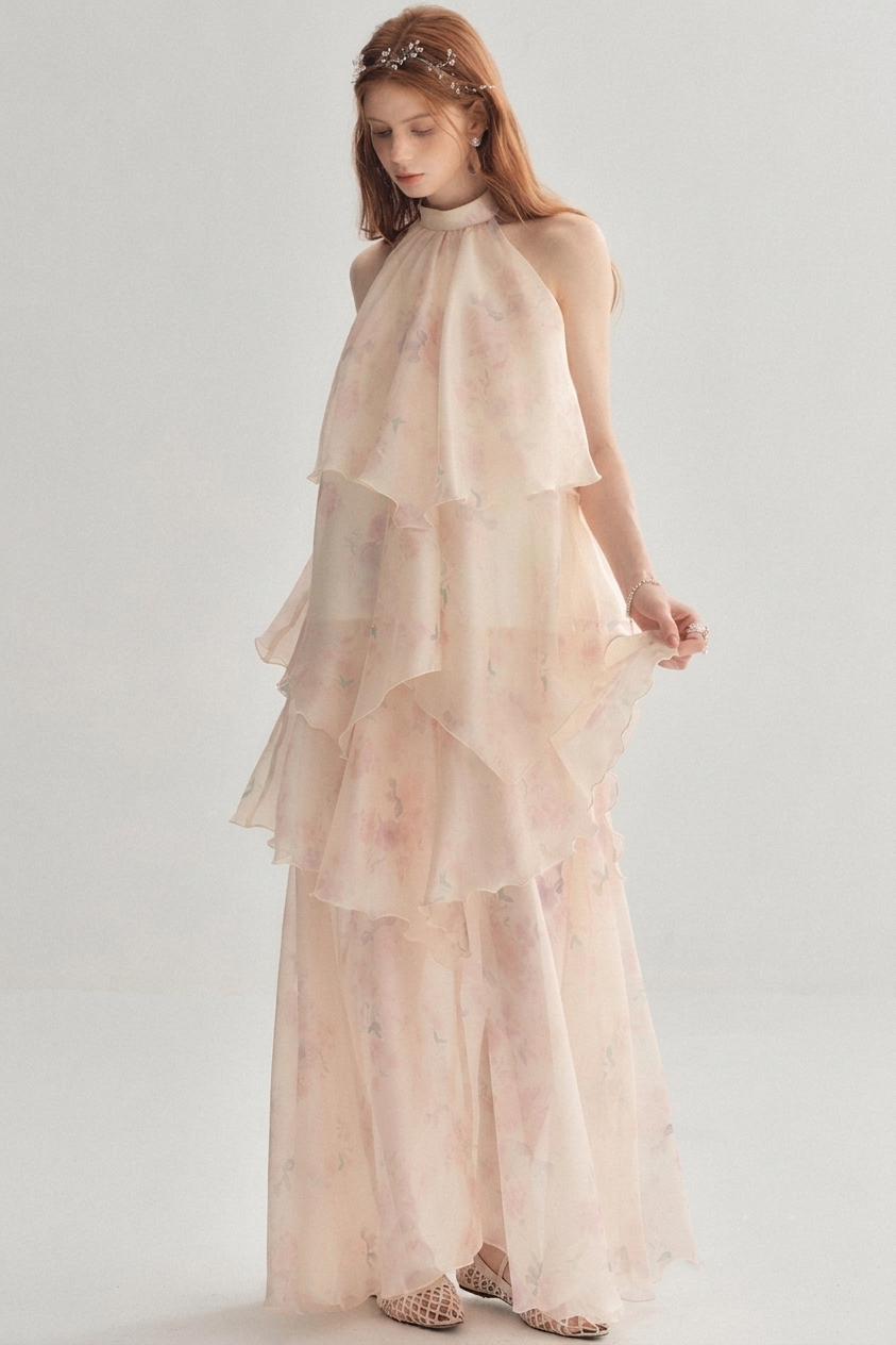 A woman modeling a sleeveless, high-neck, tiered maxi dress in a very light blush pink color with a subtle floral print, paired with a matching headpiece and patterned flat shoes, set against a plain light gray studio background.
