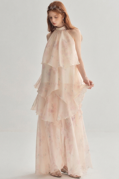 A woman modeling a sleeveless, high-neck, tiered maxi dress in a very light blush pink color with a subtle floral print, paired with a matching headpiece and patterned flat shoes, set against a plain light gray studio background.
