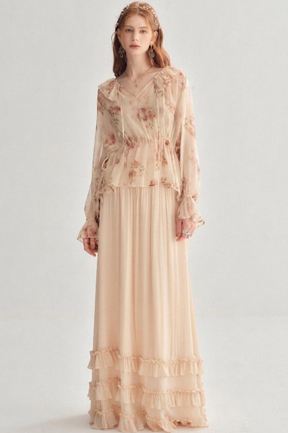 A woman modeling a long-sleeve maxi dress in a soft blush pink color with a delicate floral pattern and tiered ruffle details, set against a plain light gray studio background.