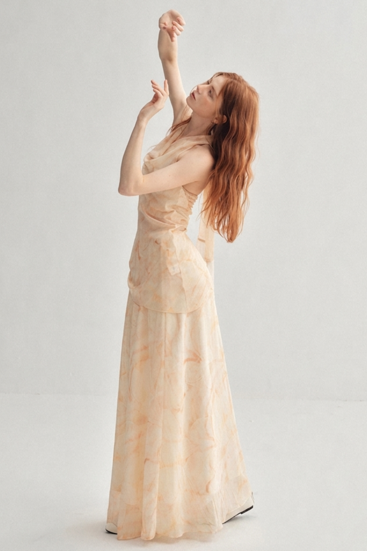 A woman with long red hair poses in a sleeveless, floor-length cream dress with a subtle floral pattern against a plain light gray studio background.