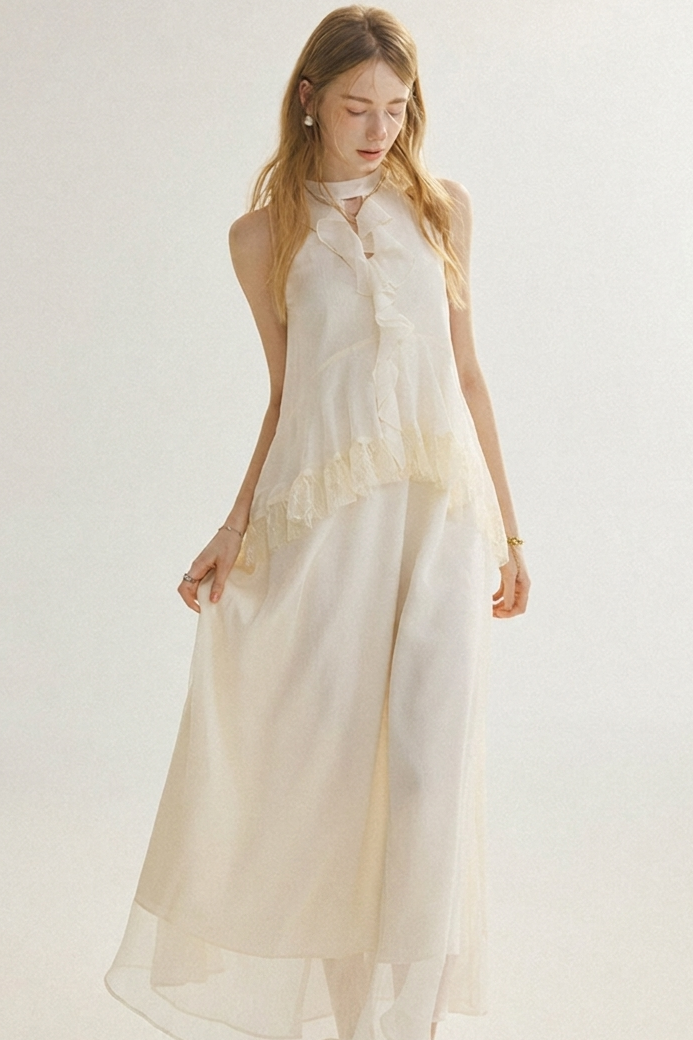 A woman modeling a sleeveless, high-neck, floor-length cream dress with layered sheer fabric and delicate lace trim, set against a plain light background.