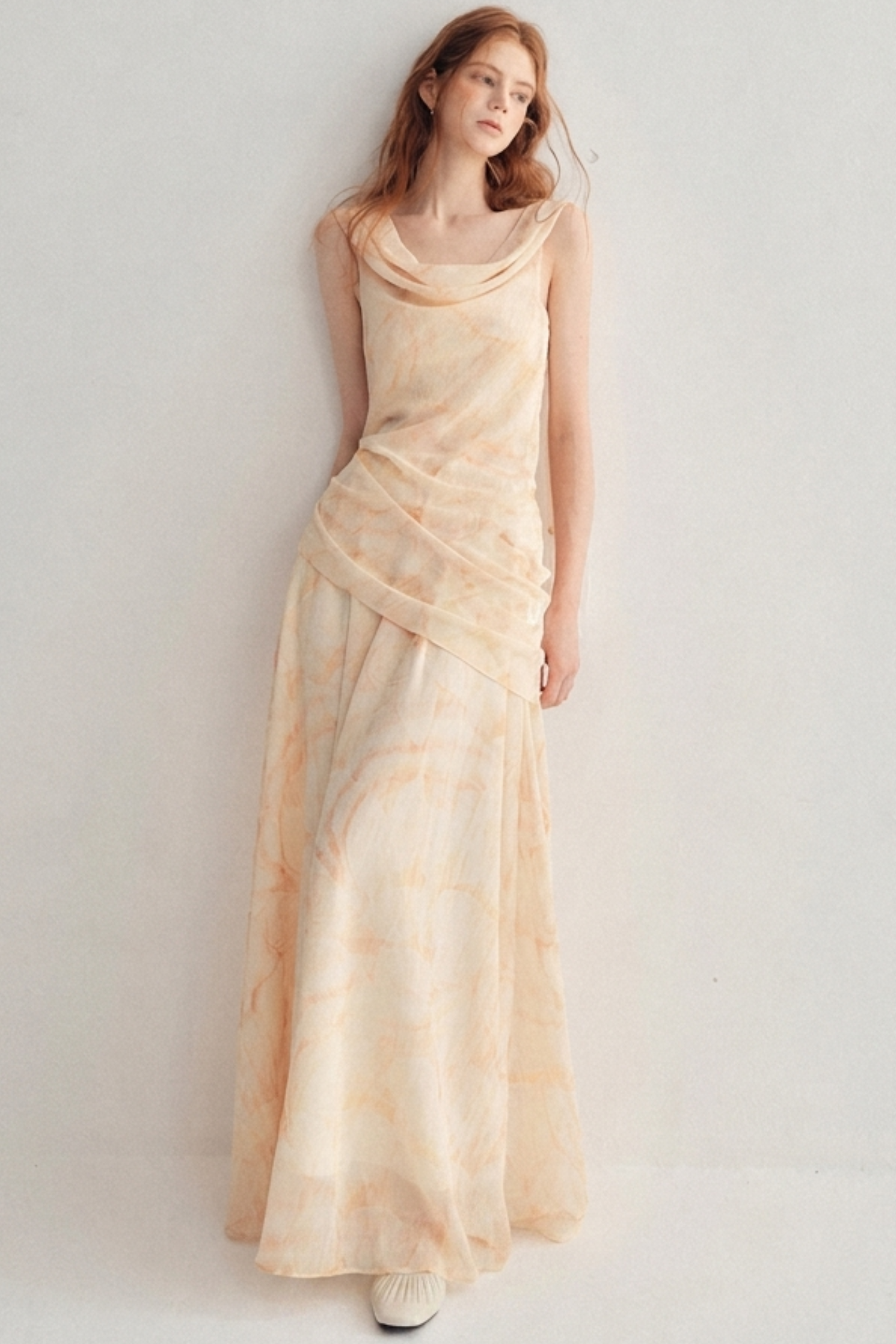 A woman wearing a sleeveless, cowl-neck maxi dress in a soft peach-beige color with a subtle tie-dye pattern, standing against a plain light gray studio background.