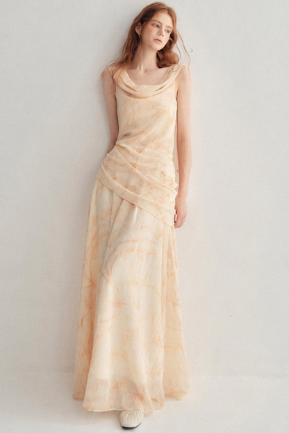 A woman wearing a sleeveless, cowl-neck maxi dress in a soft peach-beige color with a subtle tie-dye pattern, standing against a plain light gray studio background.