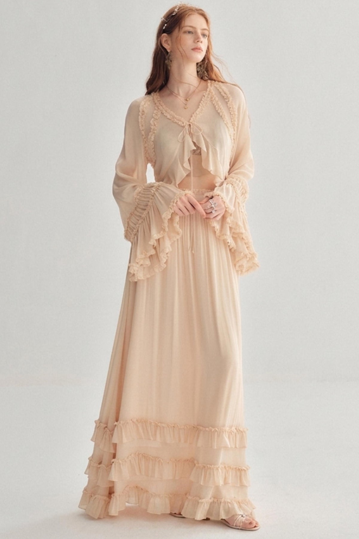 A woman modeling a long, flowing maxi dress in a soft blush beige color. The dress features a sheer long-sleeve top with ruffles, a tie at the neckline, and a tiered ruffle hem.