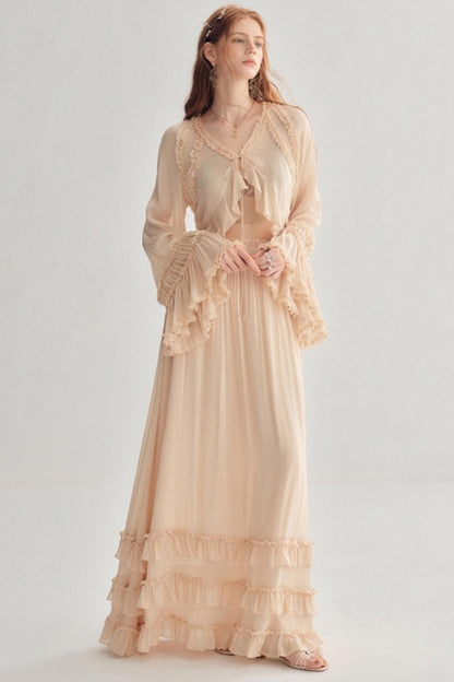 A woman modeling a long, flowing maxi dress in a soft blush beige color. The dress features a sheer long-sleeve top with ruffles, a tie at the neckline, and a tiered ruffle hem.