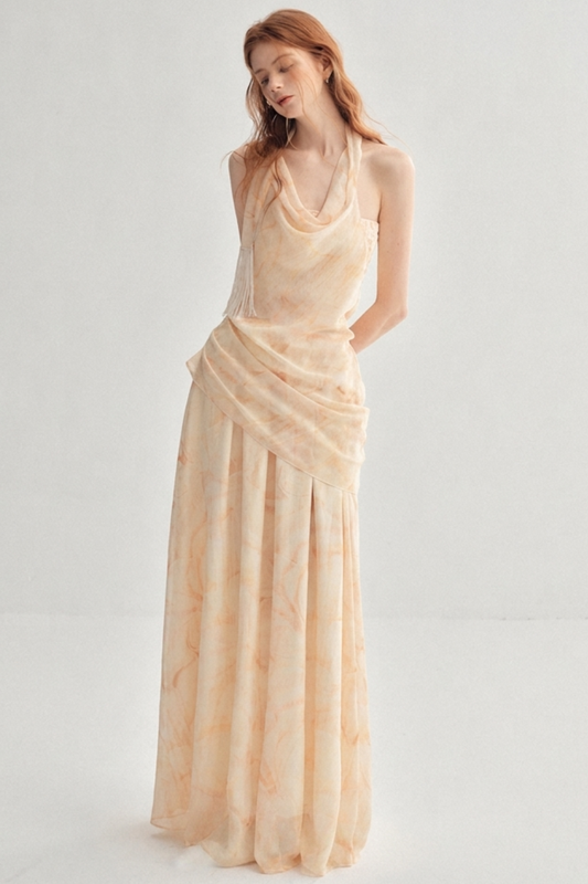 A woman modeling a sleeveless, floor-length draped gown in a soft peach-beige color with a subtle tie-dye pattern, set against a plain light gray studio background.