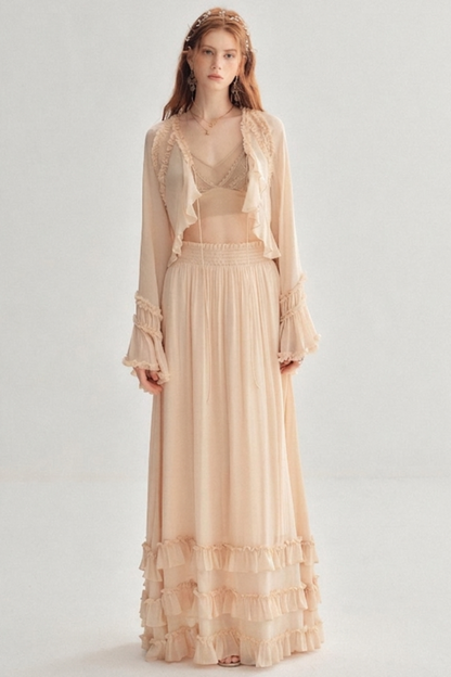 A woman modeling a light peach-beige two-piece outfit consisting of a long-sleeve sheer cropped jacket with ruffle trim over a matching crop top and a long ruffled maxi skirt, set against a plain light gray studio background.