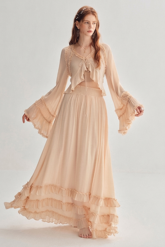 A woman modeling a long, flowing two-piece dress in a soft peach-beige color. The set features a sheer long-sleeve crop top with ruffles and a matching high-waisted maxi skirt with tiered ruffles, photographed against a light gray studio background.