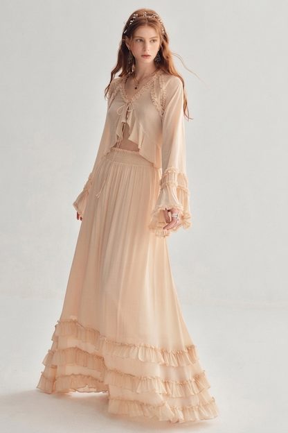 A woman modeling a light peach-beige bohemian maxi dress with a sheer long-sleeve crop top, lace details, and a tiered ruffled hem against a light gray studio background.