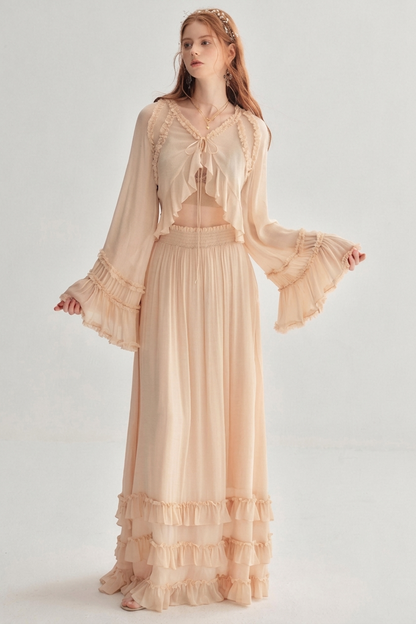 A woman modeling a light peach-beige bohemian two-piece set consisting of a long-sleeve cropped top with ruffles and a matching tiered maxi skirt, against a plain light gray background.