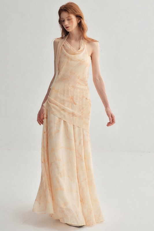 A woman modeling a sleeveless, floor-length draped dress in a soft peach-beige color with a subtle tonal floral pattern, set against a plain light gray studio background.