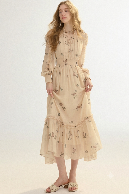 A woman modeling a long-sleeve midi dress in a light beige or cream color with a delicate floral print. The dress features a gathered waist, ruffled collar, and a tiered skirt.